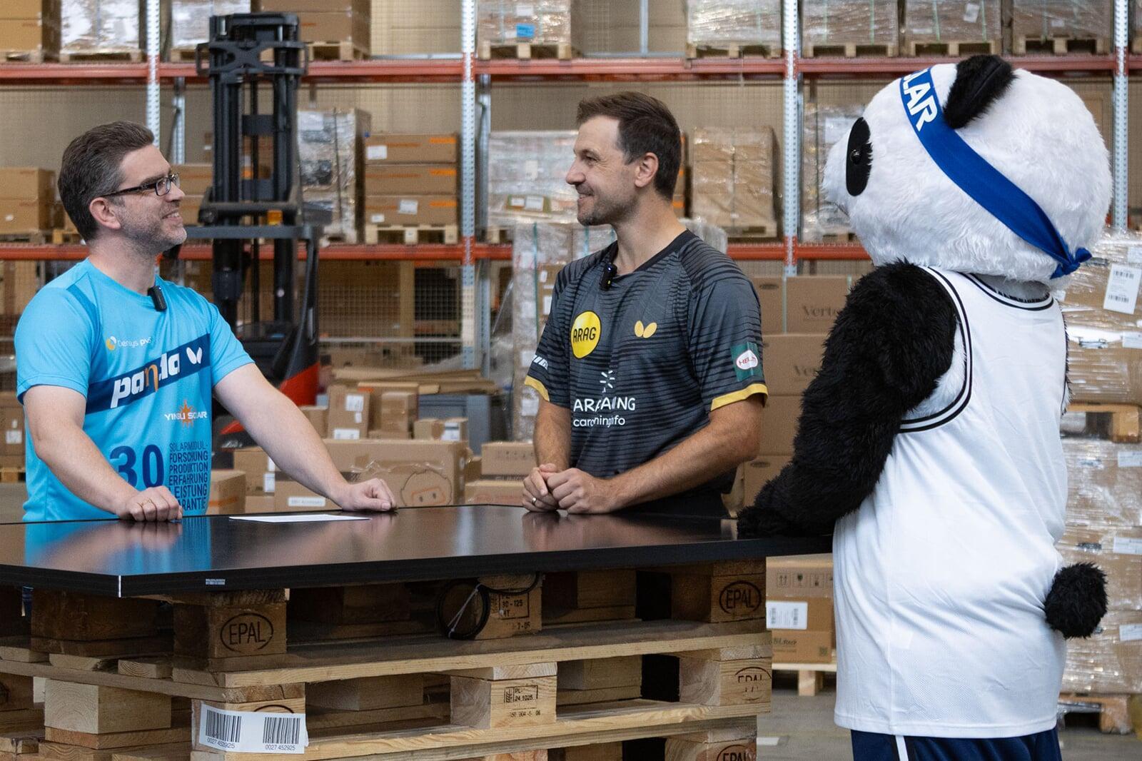 Timo Boll im Interview mit Markus Rohé und dem Panda-Maskottchen von Yingli vor einem Stapel Panda-Module Von links nach rechts: Markus Rohé, Timo Boll, Panda-Maskottchen